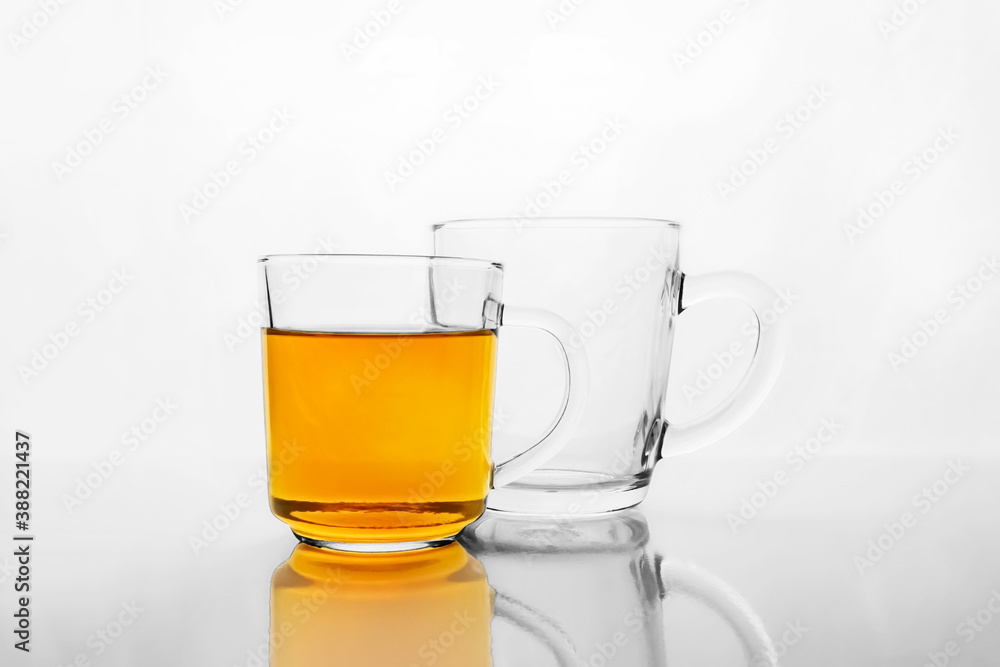 Two transparent glass mugs, one is empty and other with yellow liquid, tea or juice, on white reflective surface. Two transparent glass mugs, one is empty and other with yellow liquid, tea or juice, on white reflective surface.