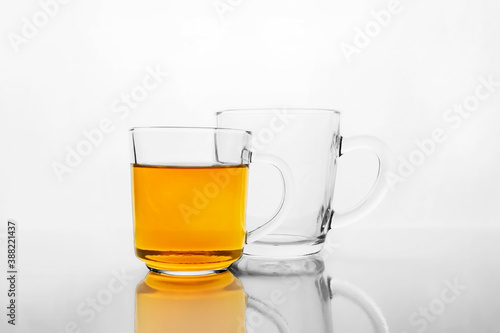 Two transparent glass mugs, one is empty and other with yellow liquid, tea or juice, on white reflective surface.