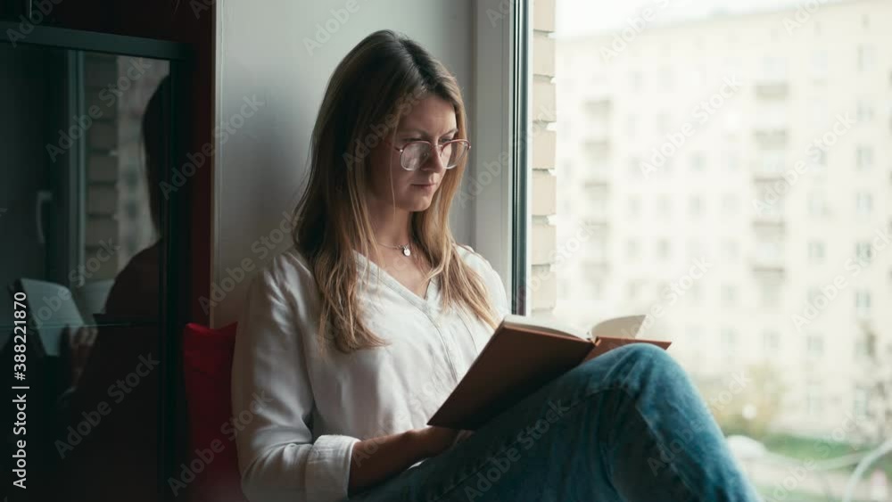 A young woman with glasses sits on the windowsill and reads a book. Cozy indoor activity. A young woman with glasses sits on the windowsill and reads a book. Cozy indoor activity.