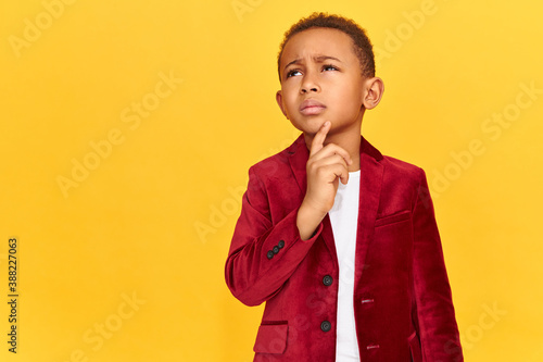 Thoughtful forgetful dark skinned schoolboy in bright velvet jacket looking up with pensive doubtful facial expression, holding fore finger on his chin, trying to remember word while reciting poem