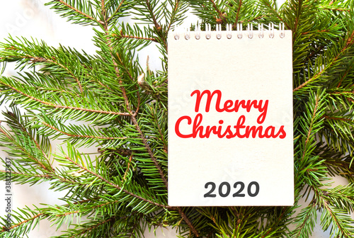 Brown notepad lying on branches of a christmas tree with text Merry Christmas 2020 © Andrey