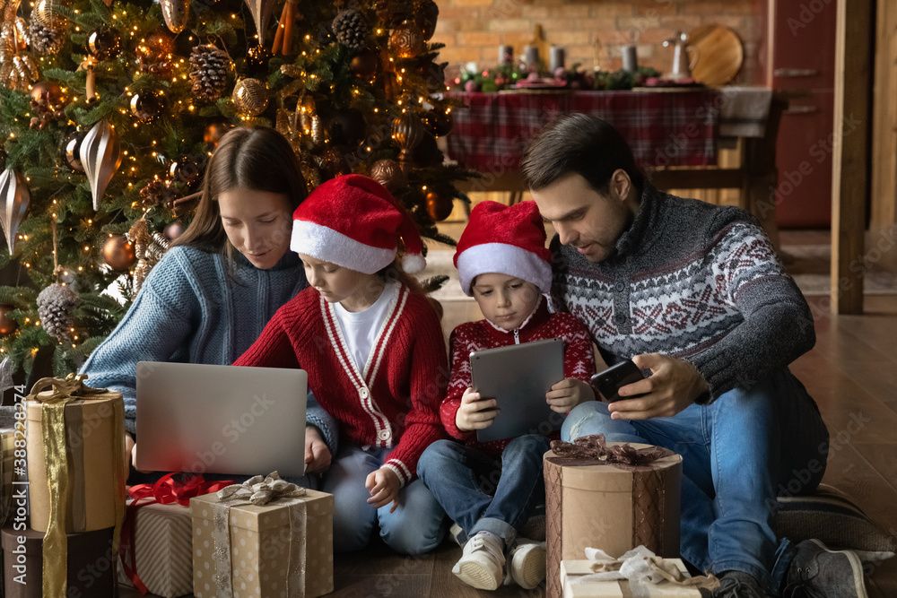 Happy young Caucasian family with small kids sit on floor near Christmas tree use gadgets together. Smiling modern parents with little children browse internet on devices, celebrate New Year at home.