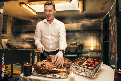 Young chef cuts fried lamb hams with a knife, in the smoky atmosphere of an old kitchen photo