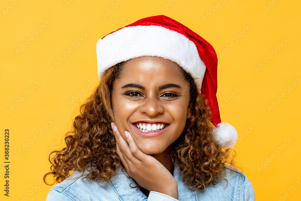 Close up portrait of amazed happy African American woman wearing Christmas hat smiling with hand holding chin on yellow isolated background Close up portrait of amazed happy African American woman wearing Christmas hat smiling with hand holding chin on yellow isolated background