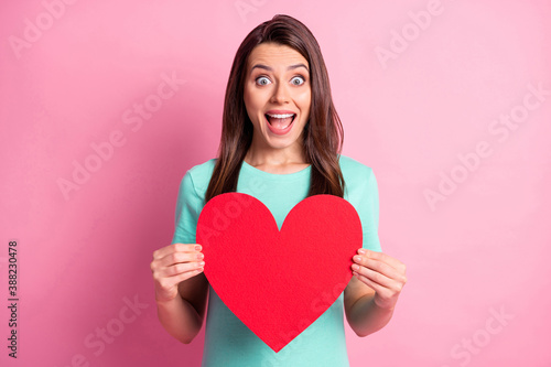 Photo portrait of amazed shocked girl keeping red heart shaped invitation card smiling staring isolated on pastel pink color background © deagreez