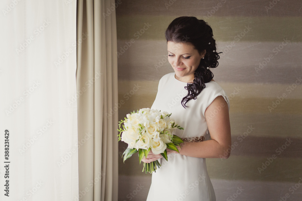 Pretty young Bride in simple wedding dress with her wedding bouquet. Black-haired woman with wedding hair-style with a long tail. Boudoir morning of the bride in modern room Pretty young Bride in simple wedding dress with her wedding bouquet. Black-haired woman with wedding hair-style with a long tail. Boudoir morning of the bride in modern room