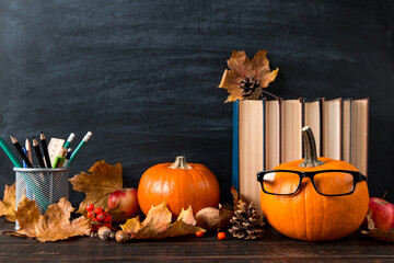 Pumpkin with glasses, halloween concept, books, pens, fallen leaves, on the table, on background of a chalkboard. Template concept of autumn mood.