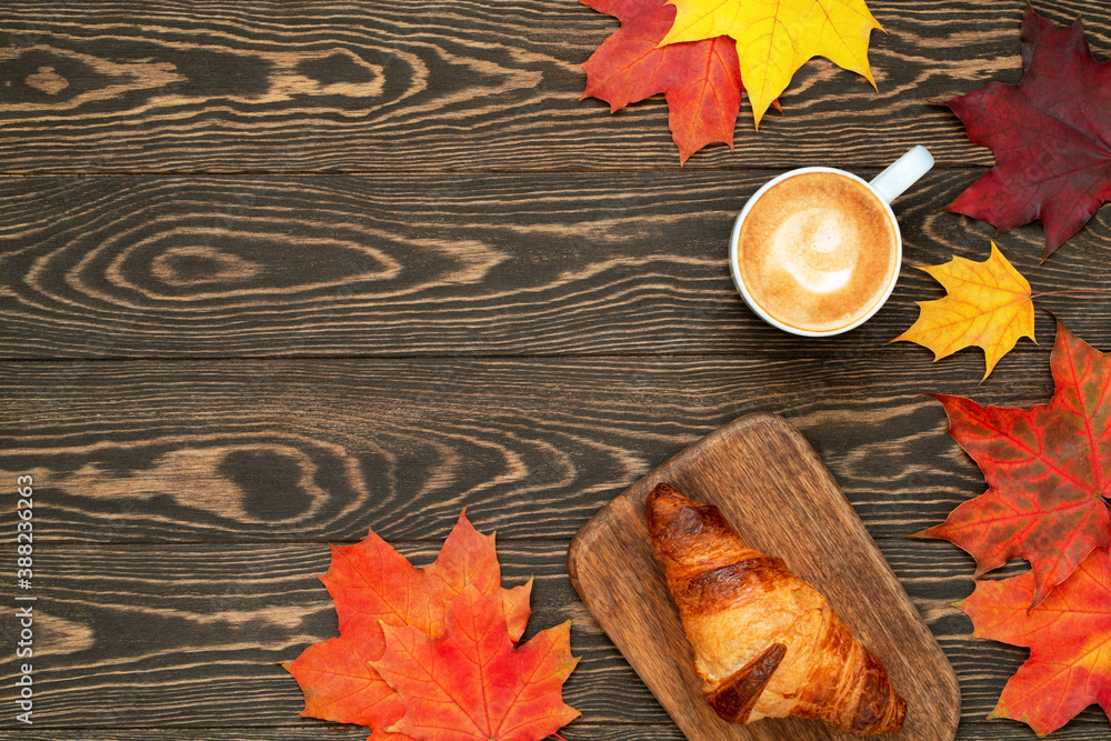 Obraz premium A cup of coffee, homemade croissant and colorful maple leaves on the wooden background. Autumn flat lay with copy space. Good morning breakfast.