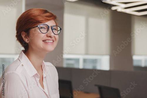Close up of a happy young woman wearing glasses, smiling joyfully at her workspace