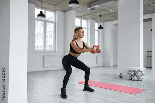blonde squats with pink dumbbells. A young sportswoman trains her legs in a light gym. healthy lifestyle