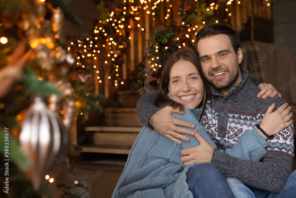 Fototapeta premium Portrait of happy young couple pose near decorated Christmas tree hugging cuddling celebrating winter holidays together. Smiling man and woman enjoy New Year celebration at home together.