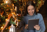 Smiling young Caucasian woman sit near decorated Christmas tree have fun browsing internet on tablet. Happy millennial female talk on video call, communicate online on webcam using pad gadget.