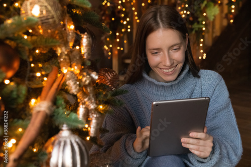 Smiling young Caucasian woman sit near decorated Christmas tree have fun browsing internet on tablet. Happy millennial female talk on video call, communicate online on webcam using pad gadget.