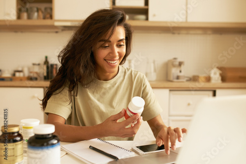 Biohacking and superfoods concept. Cheerful young Latin female nutritionist holding bottle of dietary supplements for muscle building. Pretty girl using laptop ordering vitamins via online store