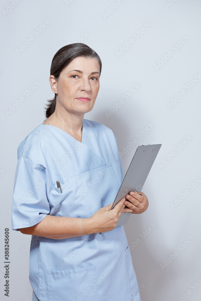 Portrait of mature female nurse practitioner in a blue scrub holding a clipboard, light background. Portrait of mature female nurse practitioner in a blue scrub holding a clipboard, light background.