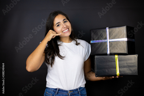 Young beautiful woman holding gifts over isolated black background smiling doing phone gesture with hand and fingers like talking on the telephone