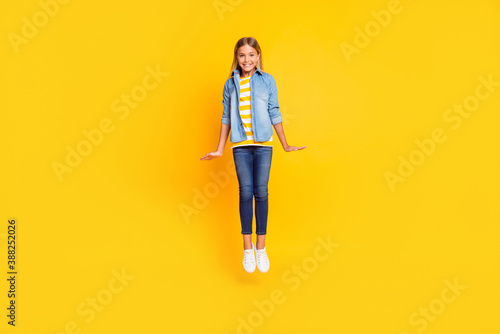 Full length body size photo of smiling shy girl with blonde hair jumping high keeping hands along body isolated on bright yellow color background