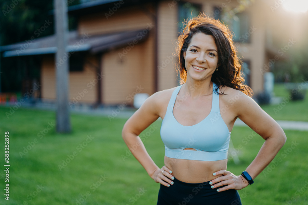 Beautiful pleased curly haired woman in good shape, keeps hands on waist, dressed in cropped top, smiles gladfully, has sporty body, poses outdoor against house background. Morning workout concept
