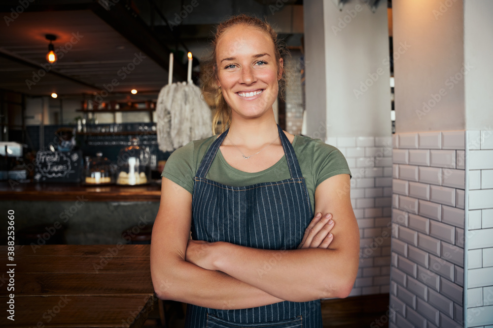 Portrait of beautiful young female waitress standing with arms crossed in cafe and wearing apron