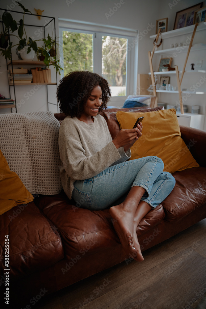 Obraz premium Beautiful african female watching video on smartphone at home while relaxing on couch