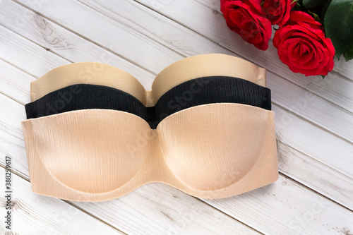 beige and black strapless nude bra on a wooden texture background next to rose flowers . Copy space for text input on health care and fitness. Balconette bra and lingerie for woman, top view.