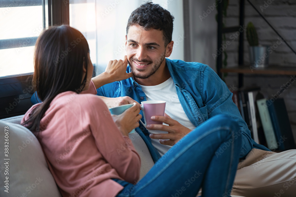 Lovely couple holding coffee cups while looking at each other and smiling sitting on couch at home. Lovely couple holding coffee cups while looking at each other and smiling sitting on couch at home.