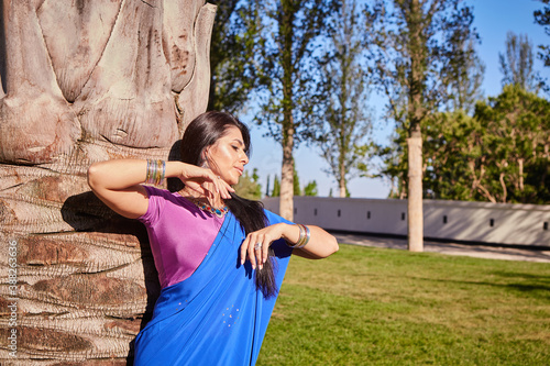 A beautiful young Indian woman in traditional clothes with makeup and jewelry stands near a palm tree. In the open air under the bright rays of the sun. Indian beauty