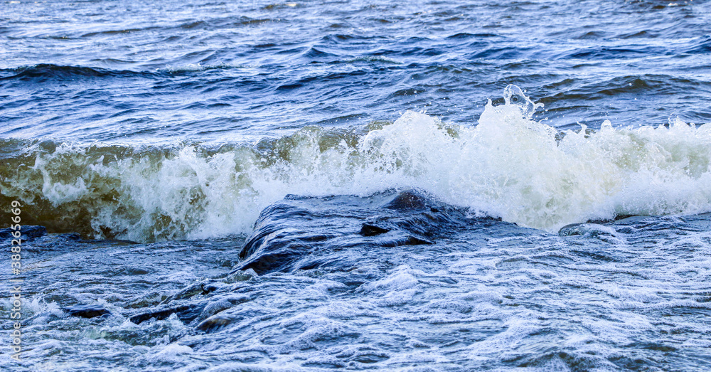 Naklejka premium waves run onto the shore and crash against the rocks, creating many splashes and splashes near the shore. river surf in stormy weather near a stone pebble coast with foamy splashing waves.