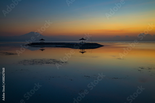 Sunrise seascape. Mountains and Agung volcano. Traditional gazebos on an artificial island in the ocean. Water reflection. Sanur beach, Bali, Indonesia.