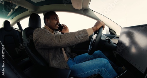 Happy young African American male traveling in self-driving electric car with autopilot and speaking on smartphone. Handsome man sitting in driverless vehicle and talking on cellphone. © VAKSMANV