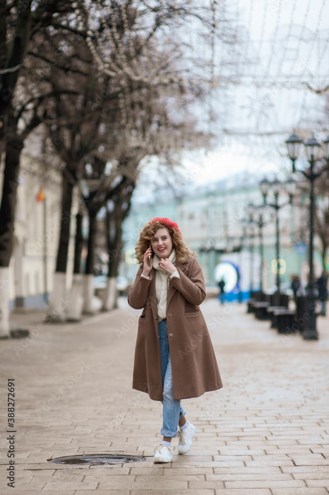 Naklejka premium A girl in a coat and a red hat is walking down the street and talking on the phone