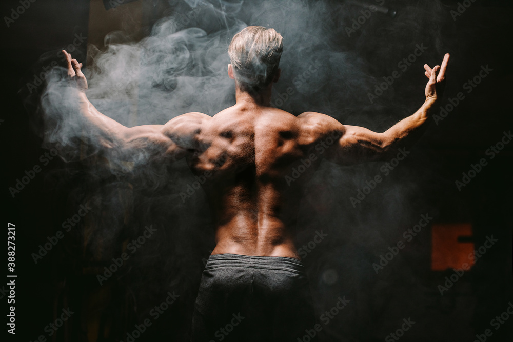 muscular man posing in gym. Fitness athlete man showing back muscles in gym. Muscular male shirtless posing with hands up. Black background.