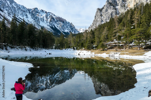 A woman standing next to a shore of Green Lake in Austrian Alps in winter and taking pictures of crystal clear water, great visibility. Reflections of Alps in calm lake's water. Winter wonderland.
