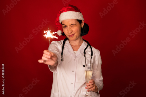 attractive sexy female doctor wink looking at camera with glass of champagne and Burning sparklers in her hands celebrating Christmas and New Year on red studio backdrop copy space