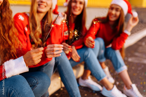 Company of cheerful young women celebrating the New Year in Santa Claus hats, with sparklers, outdoors