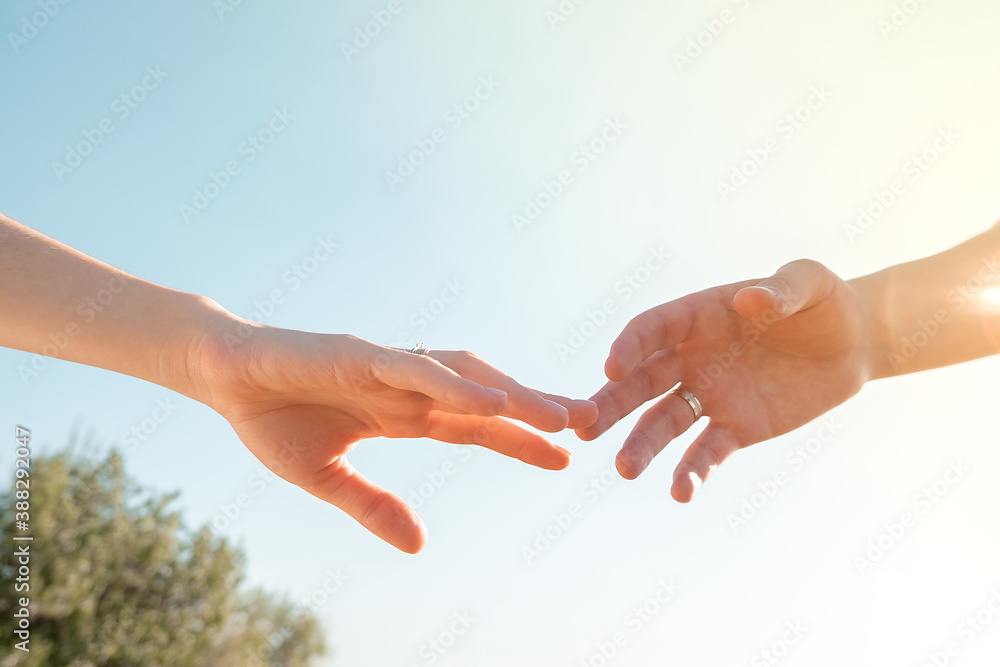 two hands with wedding rings against a blue clear sky are reaching out to each other..