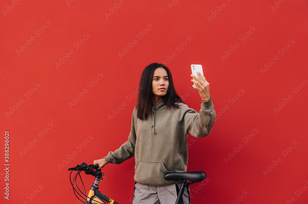 Hispanic woman standing with bicycle and taking selfie on black wall background. Lady with a bicycle takes a selfie, isolated.