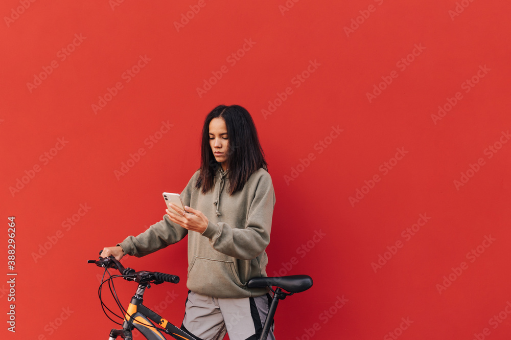 Pretty hispanic woman with bicycle in hand uses smartphone on red background. Beautiful woman with red bicycle isolated. Copy space Pretty hispanic woman with bicycle in hand uses smartphone on red background. Beautiful woman with red bicycle isolated. Copy space