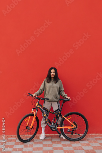 Fototapeta Naklejka Na Ścianę i Meble -  Cheerful woman in sportswear stands with a bicycle on a background of a red wall and poses for the camera. Cycling down the street.