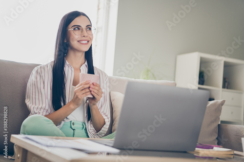 Portrait photo of dreamy cheerful young woman keeping cup of coffee working remotely from home sitting near laptop smiling