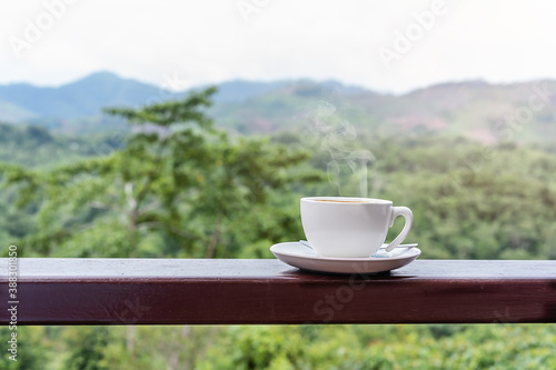 Close-up of a hot white coffee cup on wooden table with the scenic view of mountain in the morning sunlight and beautiful green nature background with copy space for text