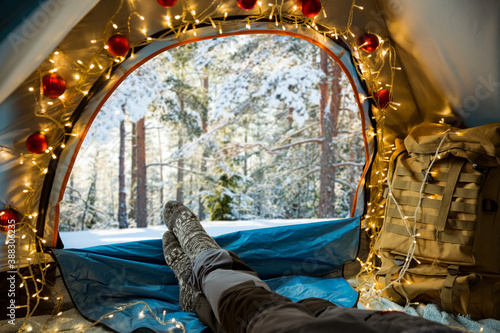A man lying in a tent decorated with Christmas lights wearing warm wooden. Beautiful winter wild forest covered with snow. Self-isolation and social distancing during holidays.