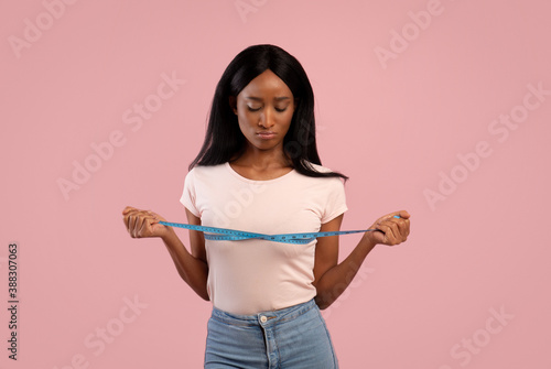 Weight loss and dieting. Upset African American woman measuring her breasts with tape on pink studio background © Prostock-studio