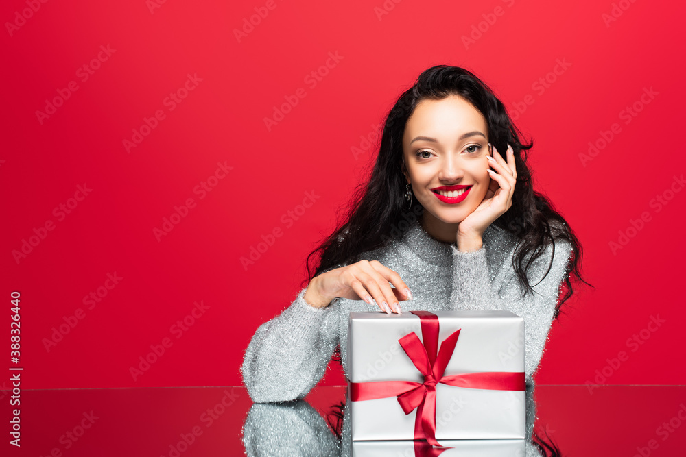 young happy woman in sweater looking at camera near wrapped present isolated on red young happy woman in sweater looking at camera near wrapped present isolated on red