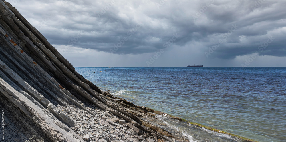 Panorama of a cloudy seascape. A picturesque stone wild beach at the foot of the rocks in rainy weather and a ship on the horizon. Surroundings of the resort of Gelendzhik. Russia, Black sea coast
