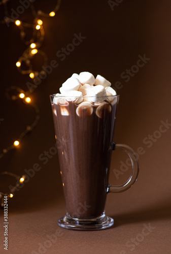 Cocoa drink with marshmallows in a glass on a brown background with festive winter decorations. Winter mood. Minimalistic monochrome composition. Selective focus. Frontal view. Vertical. Copy space