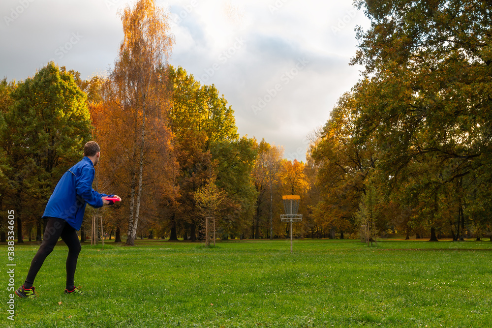 Young caucasian man in blue jacket playing disc golf on autumn play course with basket - obrazy, fototapety, plakaty 