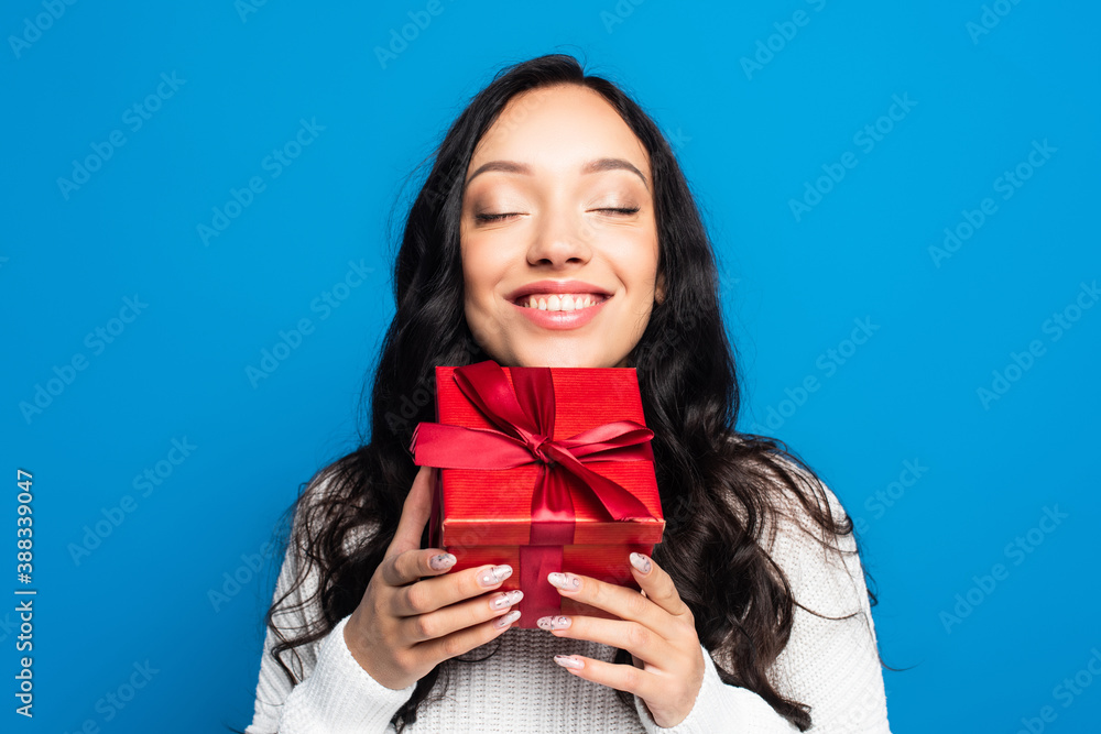 Fototapeta premium happy brunette woman with closed eyes holding christmas present isolated on blue