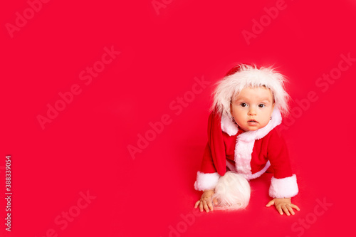 a surprised little child six months old in a Santa costume sits on a red isolated photophone, a place for text, a new year and holiday concept © Any Grant