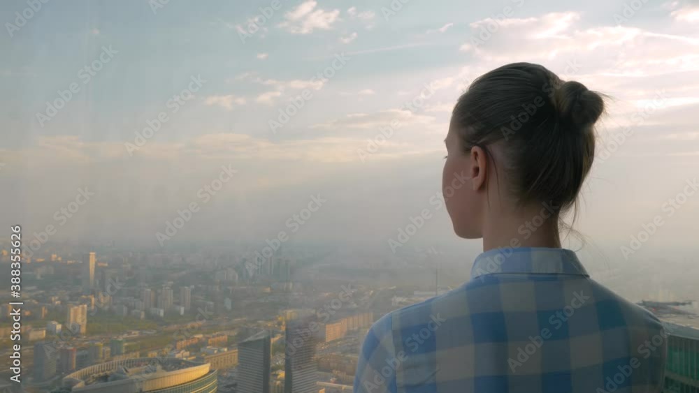 Back view of woman looking at cityscape through window of skyscraper. Summer time, warm sunset sky. Success, corporate, opportunity, sightseeing, discover and future concept Back view of woman looking at cityscape through window of skyscraper. Summer time, warm sunset sky. Success, corporate, opportunity, sightseeing, discover and future concept